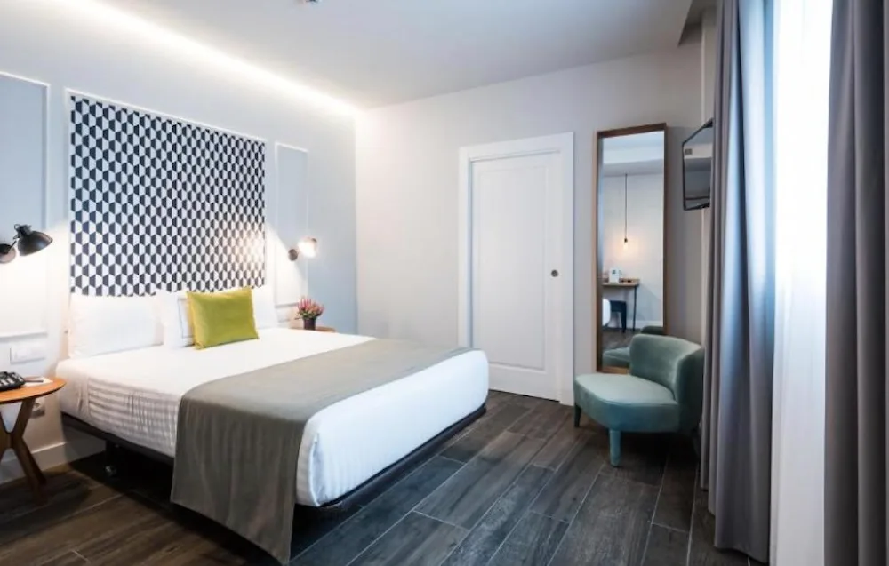 Pale blue bedroom with a black and white chequered headboard panel, a yellow cushion on the bed, a teal armchair and a tall mirror by the doorway.