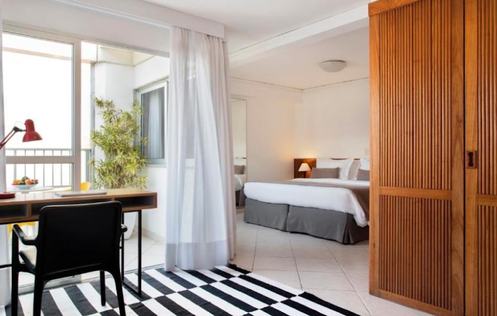 Bedroom seen through a wooden sliding door, with a black and white striped rug, a desk by the window and glass doors onto a planted balcony