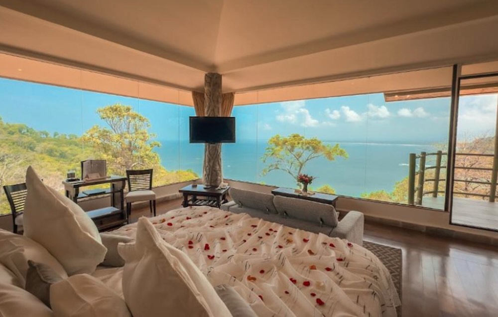 Luxury suite with floor-to-ceiling glass on three sides, a bed scattered with rose petals and wide views over the Pacific