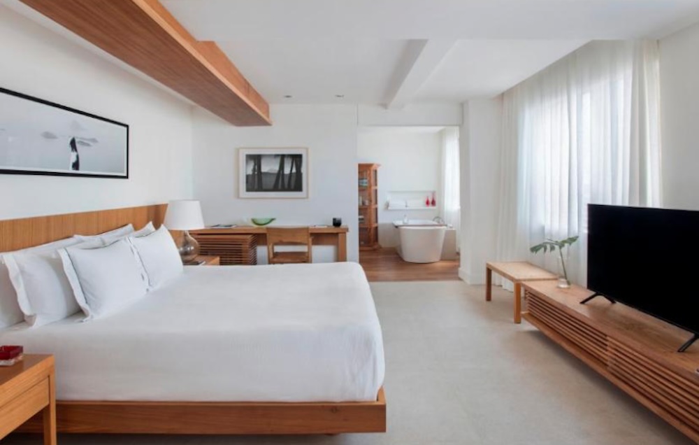 Bright, minimal bedroom in pale wood and white, with a low bed, a long desk, framed black and white photographs and a freestanding bath beyond