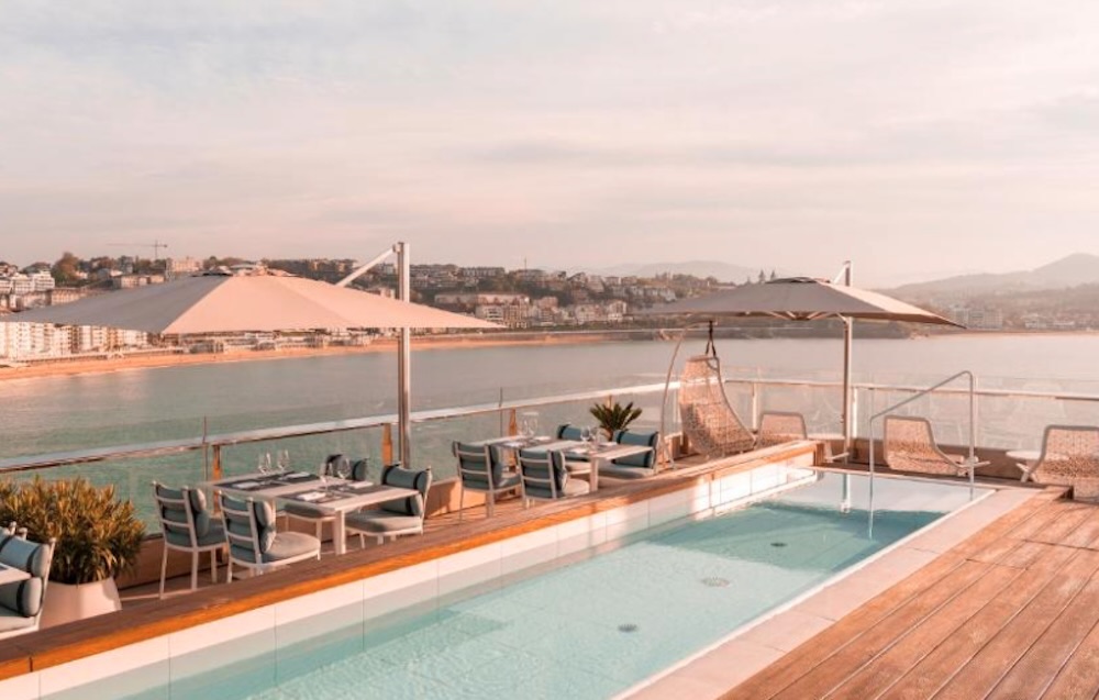 Rooftop pool and deck at sunset with parasols over set dining tables, a hanging chair at the rail and the bay curving round to the beach beyond.