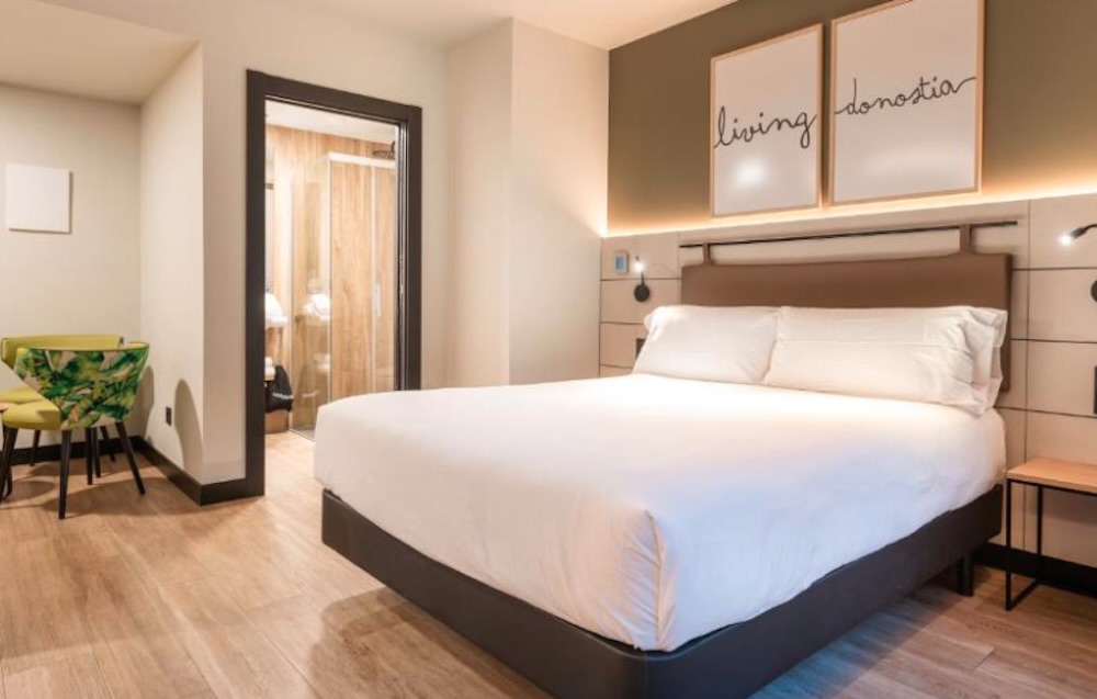 Wide white bed below two framed script artworks, a glass walled bathroom to one side and a small patterned armchair beside a round wooden table.