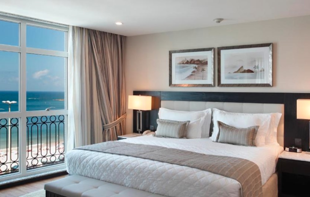 Bedroom with a large bed and framed prints, and a full-height window opening onto a balcony with a view over the beach and sea below