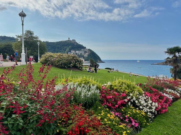 Banked flowerbeds of red, pink, white and yellow blooms with hydrangeas and lawn behind, sloping down to blue sea with a wooded headland and hilltop tower in the distance.