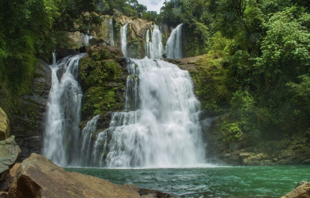 Nauyaca Waterfall in Costa Rica, water tumbling over tiers of dark rock into a green swimming pool surrounded by rainforest