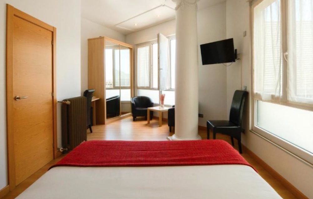 Compact white room with a structural pillar, red throw across the bed, wall mounted television and two black armchairs by tall open shutters.