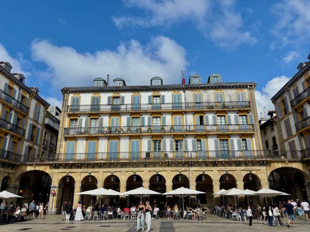 A four-storey building with rows of blue-shuttered windows and iron balconies, each numbered in yellow, above a stone arcade with café tables and parasols below.