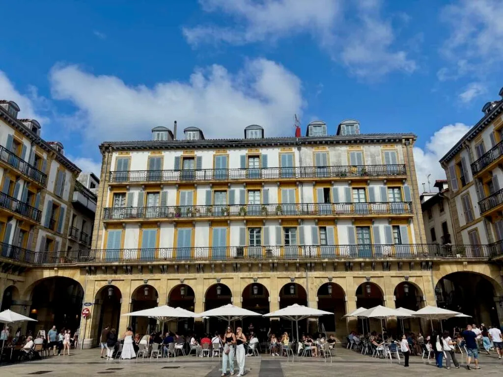 A four-storey building with rows of blue-shuttered windows and iron balconies, each numbered in yellow, above a stone arcade with café tables and parasols below.