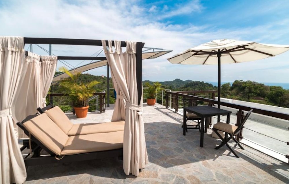 Curtained double daybed and a shaded table on a stone terrace, looking out over rainforest to the sea