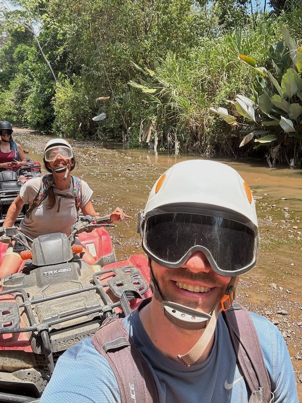 Selfie in helmet and goggles on an ATV tour near Manuel Antonio, riding through a shallow river crossing with a second rider behind