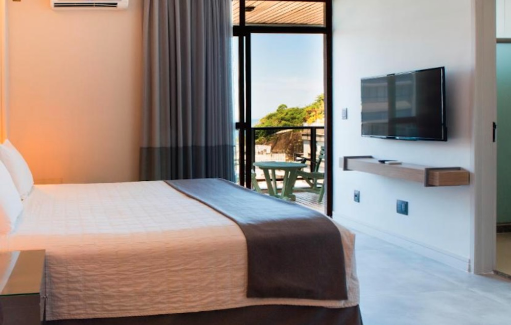 Modern bedroom with a large bed, wall-mounted television and open glass doors onto a small balcony with a table, greenery and rock visible outside