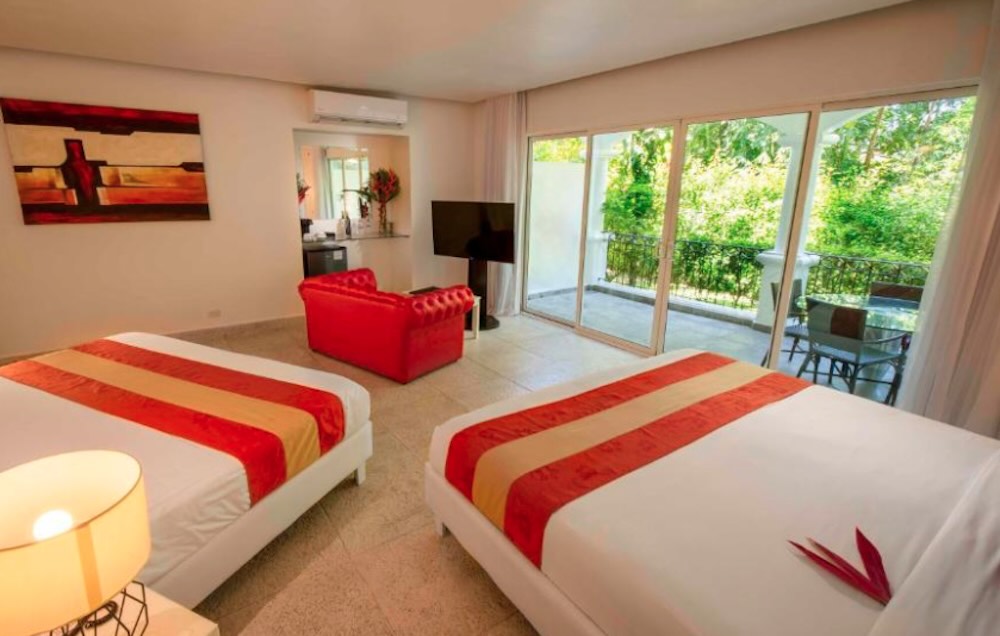 Spacious twin room with red and orange bed runners, a red leather armchair and sliding doors onto a rainforest balcony