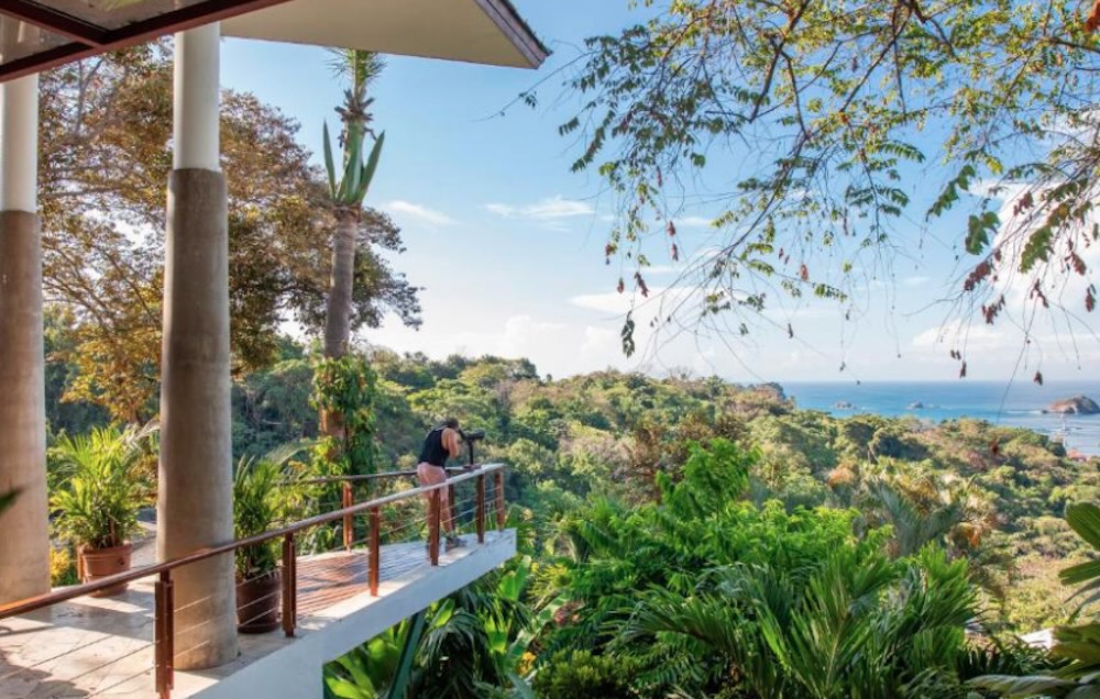 Guest using a spotting scope on a viewing deck above dense rainforest, with the ocean and an island in the distance