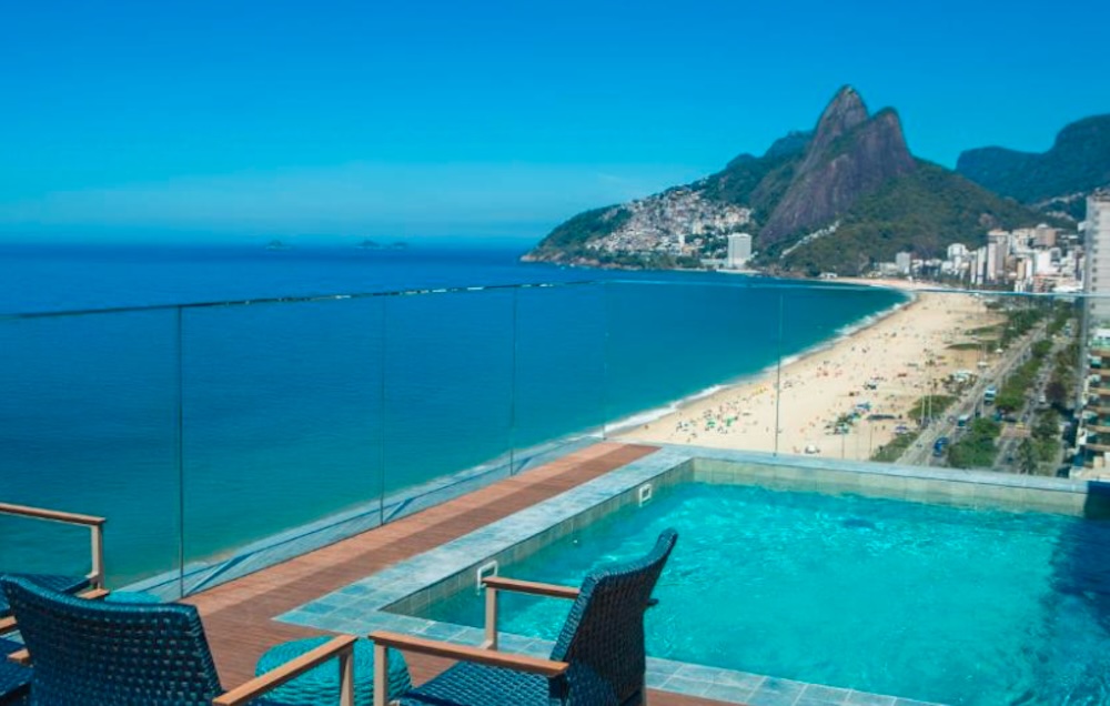 Rooftop pool with wicker chairs behind a glass balustrade, looking along a long curving beach towards the twin peaks of Dois Irmãos