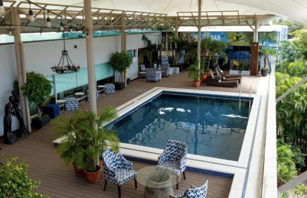 Covered rooftop terrace with a rectangular pool, timber decking, patterned armchairs and potted palms under a white canopy strung with lights