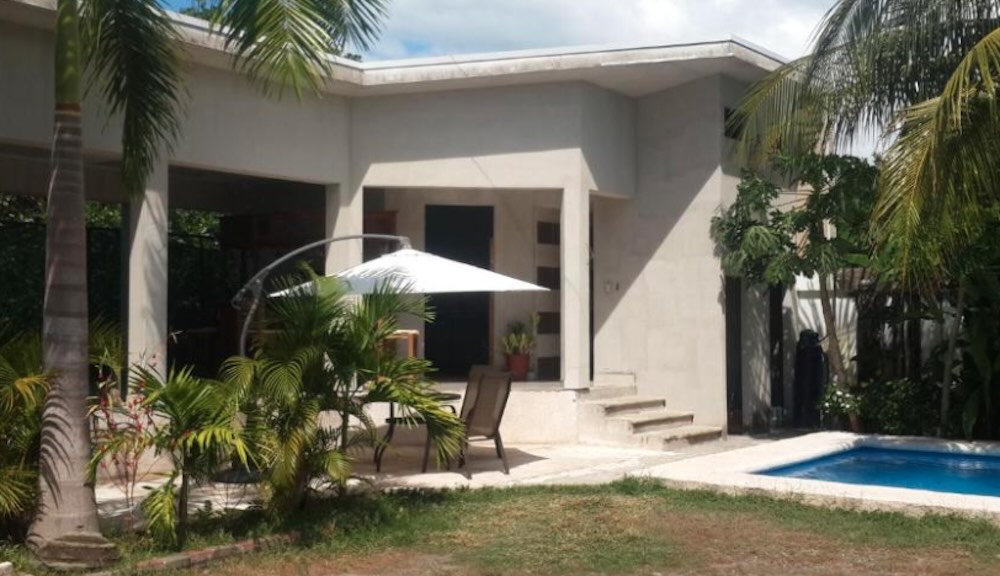 Small pool and shaded patio with a white parasol outside a low white villa, surrounded by palms and tropical planting