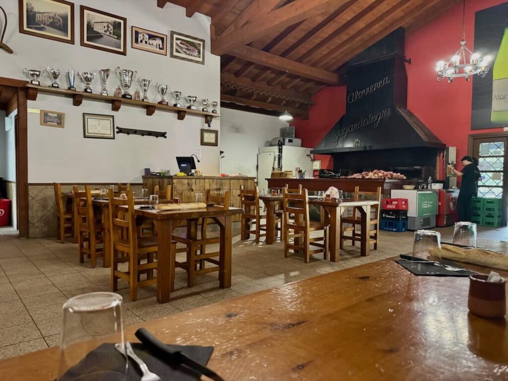Cider house dining room with long wooden tables and bread laid out, and a pile of raw txuleta stacked beside the charcoal grill.