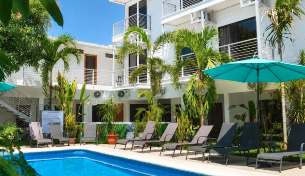 Palm-lined pool with sun loungers and a turquoise parasol in front of a white three-storey building with balconies