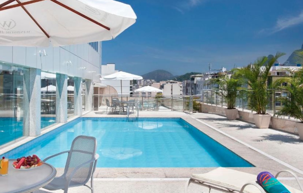 Rectangular rooftop pool edged with glass panels and white parasols, with potted palms along one side and green hills behind the surrounding rooftops