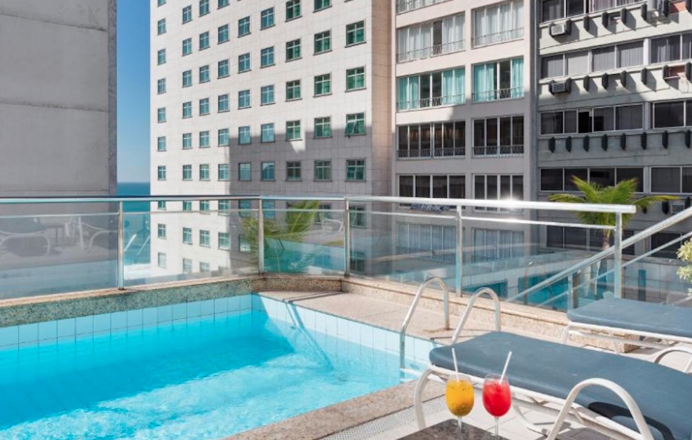 Small rooftop pool with blue sun loungers and two cocktails on a table, glass balustrade and a narrow slice of sea visible between tall buildings