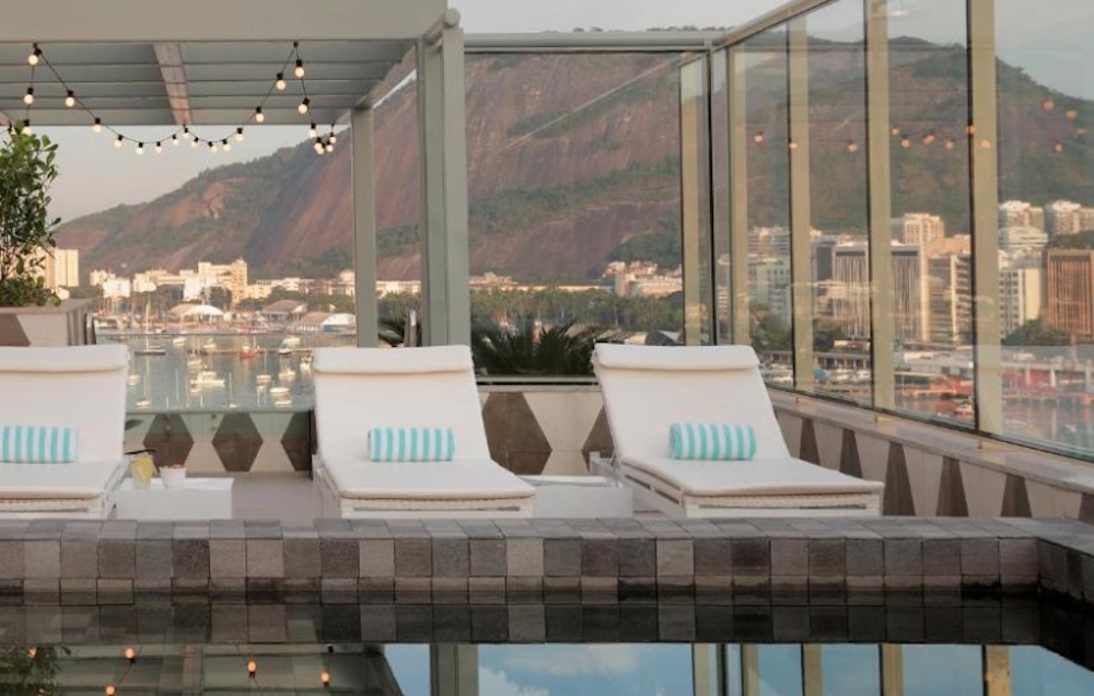 Rooftop pool with white sun loungers and festoon lights, looking out through glass to a marina, city buildings and a steep forested mountain