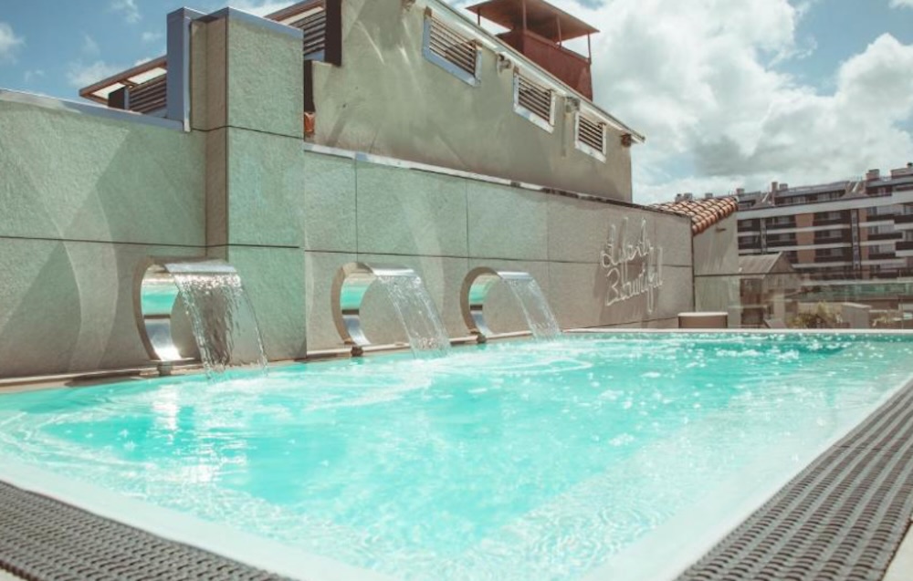 Turquoise rooftop pool with three curved chrome spouts pouring from a stone wall, grey decking around the edge and city rooftops under a cloudy sky.