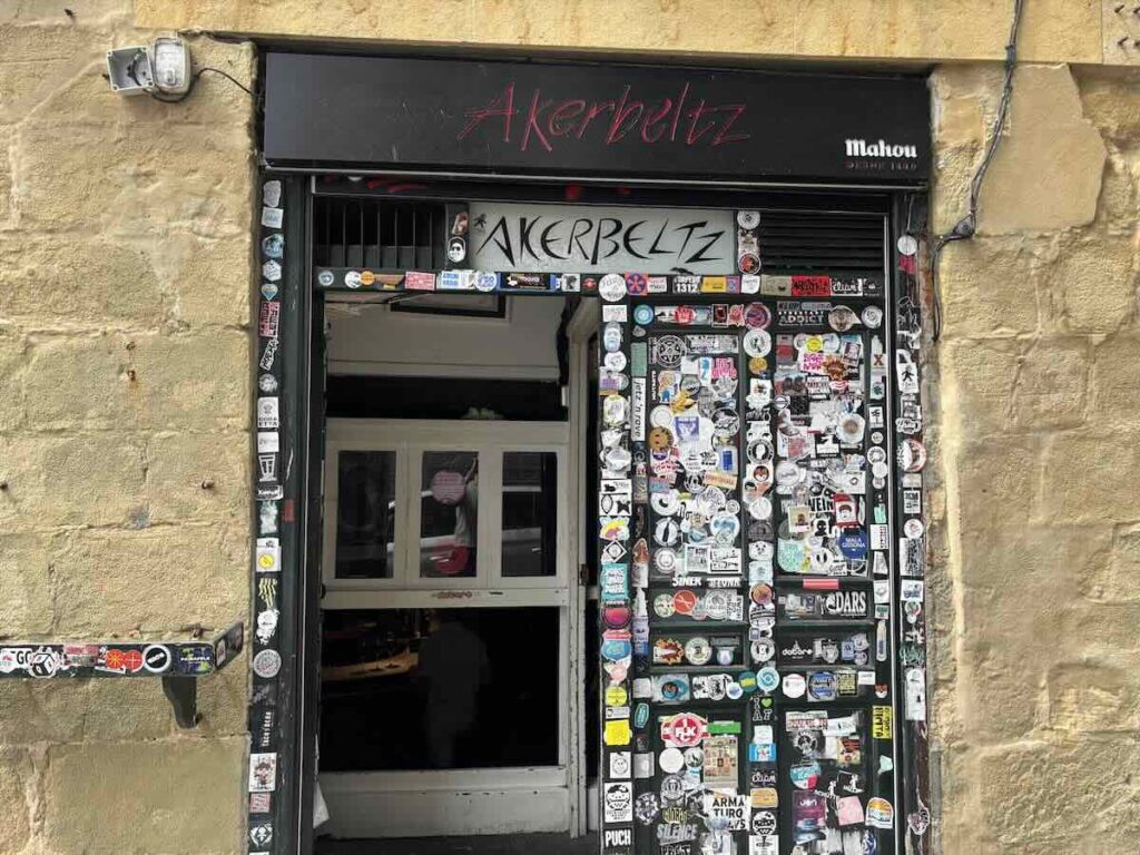 A narrow bar doorway in a sandstone wall, its frame and shutter covered edge to edge in hundreds of stickers, with the name written above in red.