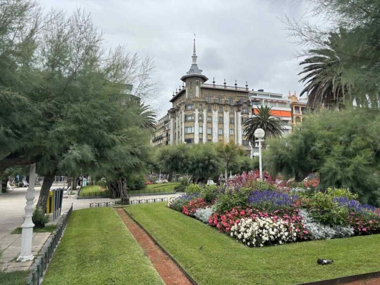 Curving lawns and dense pink, white and purple flowerbeds framed by feathery tamarisk trees, with an ornate spired building behind.