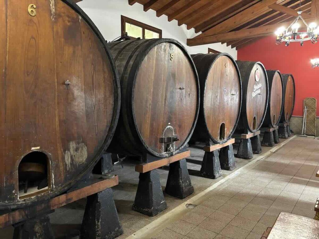 A row of six numbered wooden cider barrels on black plinths in a tiled room with exposed timber beams and a red wall.