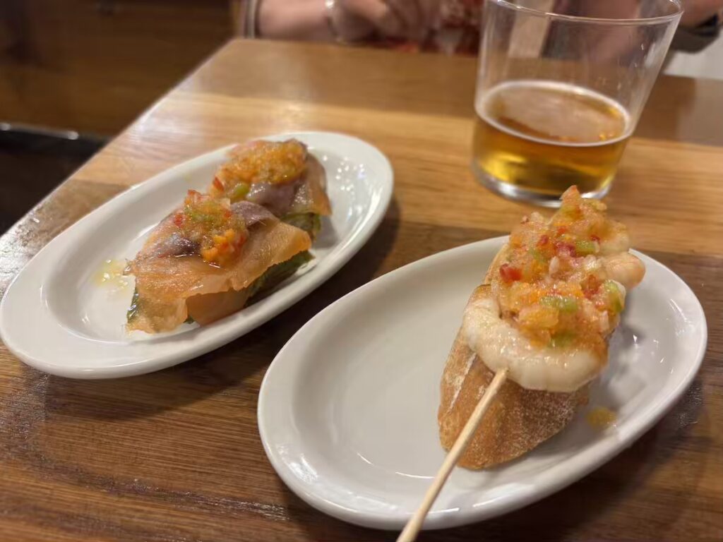 Two pintxos on small white plates, one anchovy and one prawn, each topped with chopped pepper, beside a glass of beer