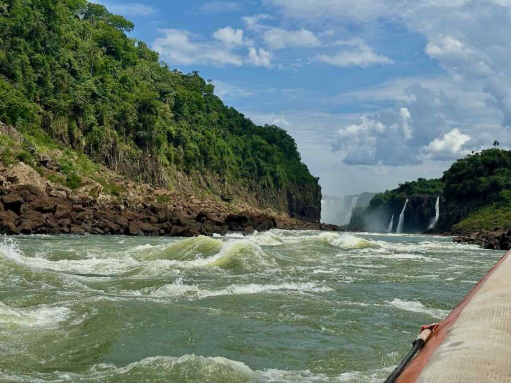View from an inflatable boat approaching white water rapids, with cliffs on both sides and waterfalls visible in the distance ahead.