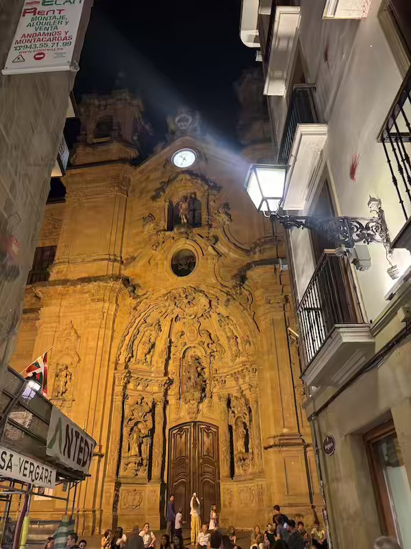 The floodlit front of the Basilica of Santa María del Coro at night, with people gathered on the steps below