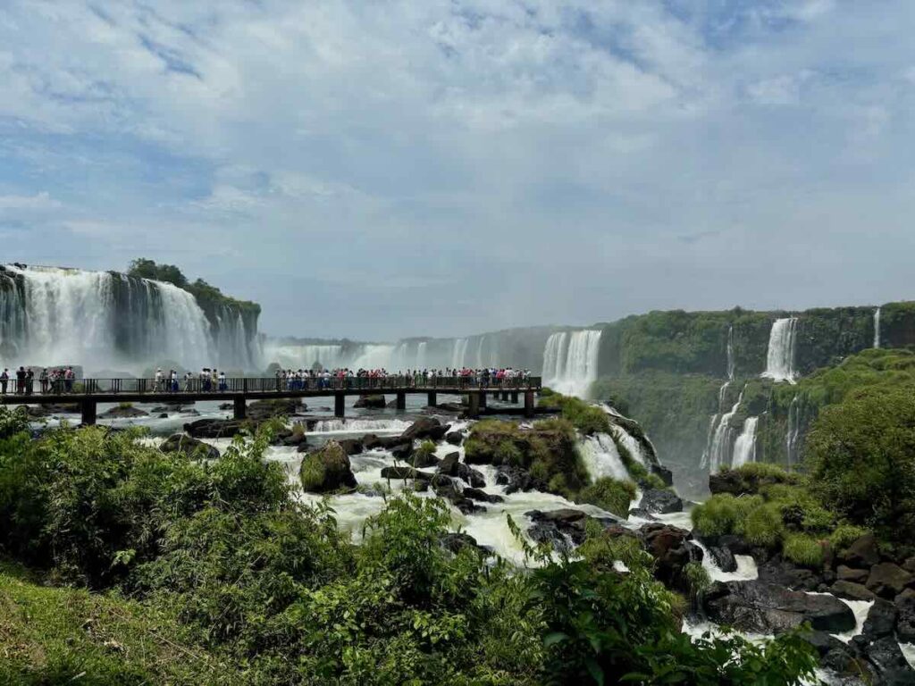 Visitors lining a low wooden walkway that crosses the river in front of a broad line of waterfalls and forested cliffs.