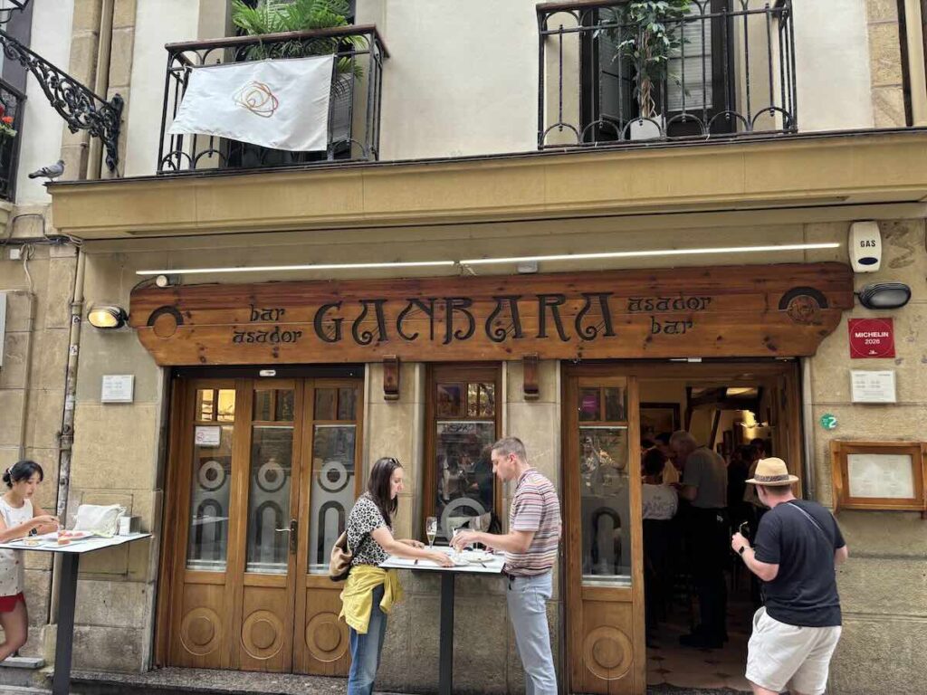 Exterior of Bar Ganbara, carved wooden sign above the door and two people eating pintxos at a table outside