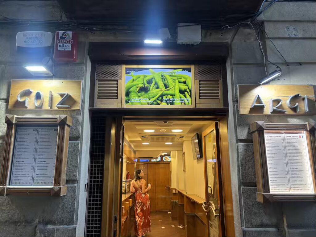 Entrance to Bar Goiz-Argi at night, printed menu boards either side and a lit sign for fried padrón peppers