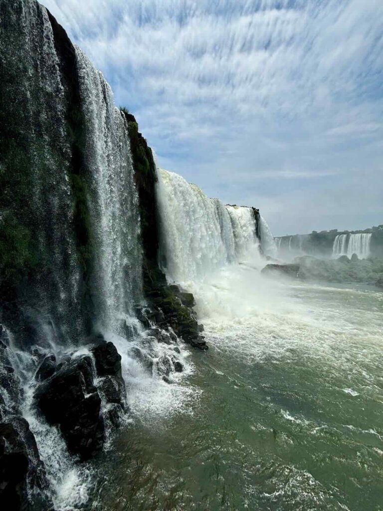 Close side view along a line of waterfalls dropping over dark basalt cliffs into a fast-moving green river, with mist rising downstream.