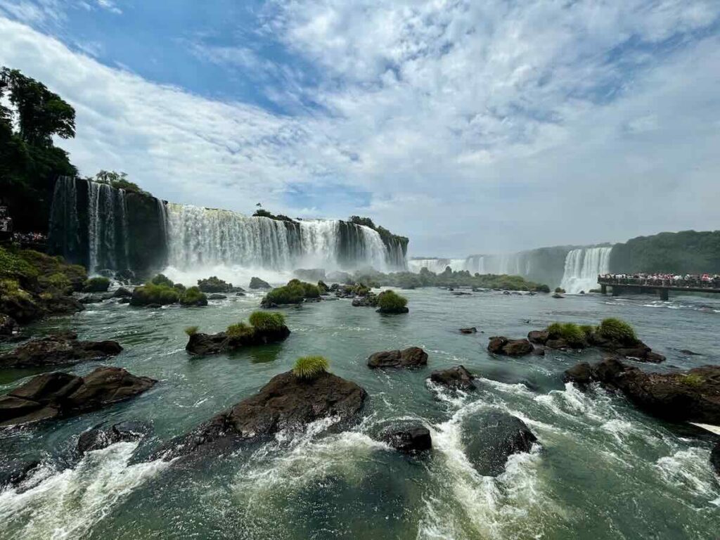 Waterfalls curving around a green gorge seen from river level, with a crowded walkway visible on the far right of the frame.