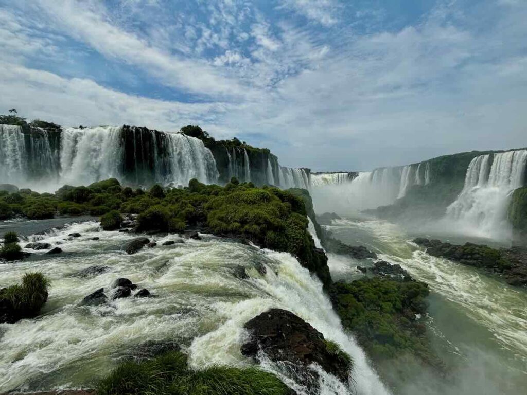 Wide view of multiple tiers of waterfalls across a green gorge, with rapids and moss-covered rocks in the foreground under a blue sky.