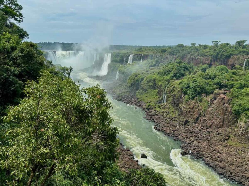 Looking down a green river gorge from a high viewpoint, with waterfalls at the far end and mist rising above the treeline.