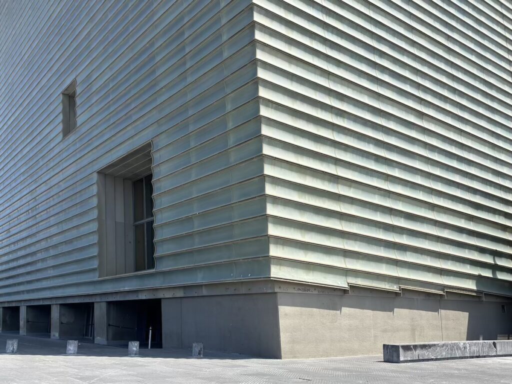 The corner of a large modern building clad in horizontal ridged translucent glass panels, with a recessed doorway and open bays at ground level.