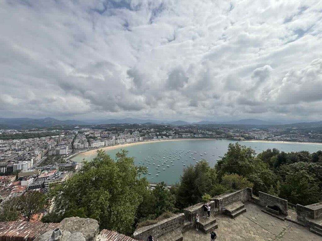 A wide bay of moored boats and a curved sand beach seen from a stone castle terrace, under heavy grey cloud with mountains beyond.