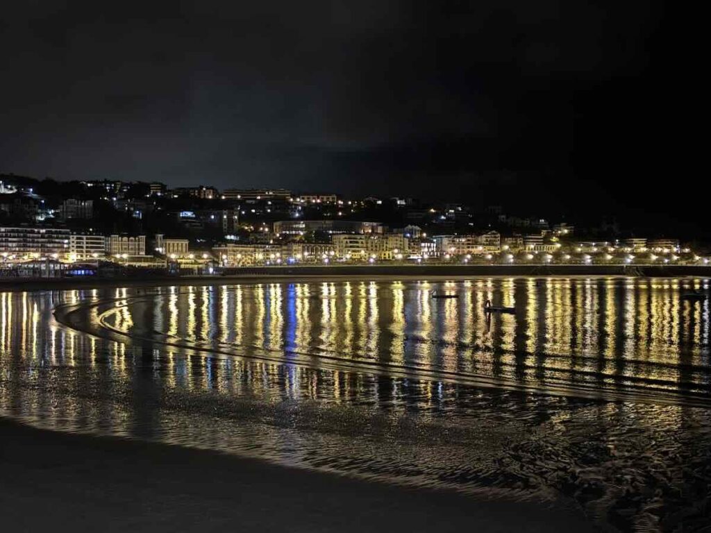 Promenade lights reflected in long streaks across wet sand at low tide, with the lit hillside of the town behind.