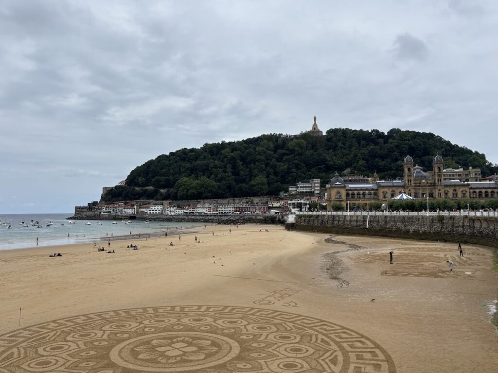A large circular geometric pattern raked into wet sand in the foreground, with the beach, a wooded hill and a domed sandstone building behind.