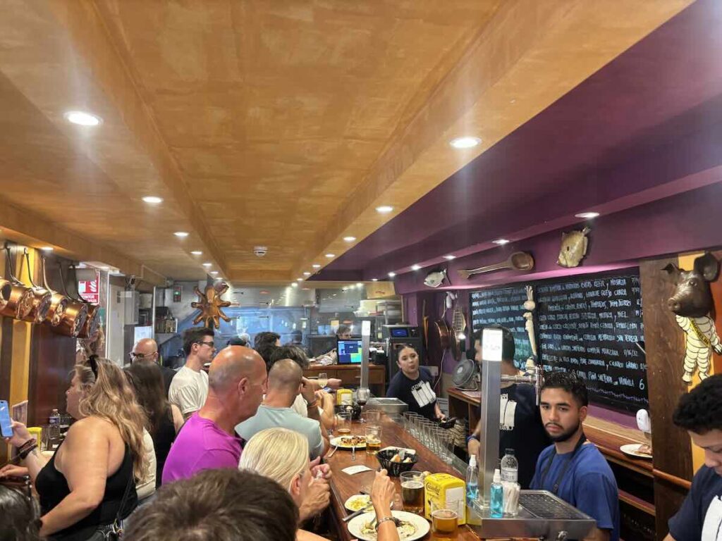 Customers packed along the bar inside La Cuchara de San Telmo, with the chalkboard menu behind