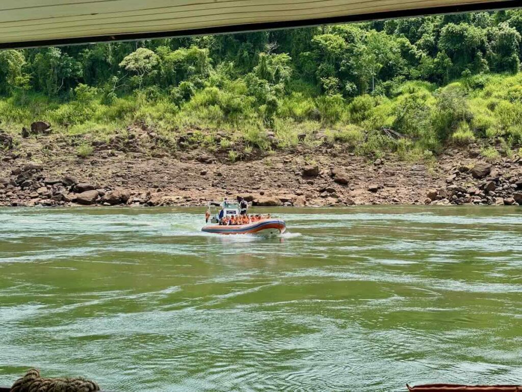 An orange and blue inflatable boat carrying passengers in life jackets along a wide green river below a rocky, forested bank.