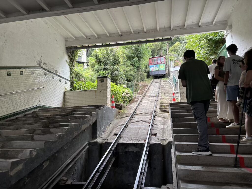 Looking up a single funicular track between two flights of stone steps, a carriage descending through greenery ahead, passengers waiting on the right.