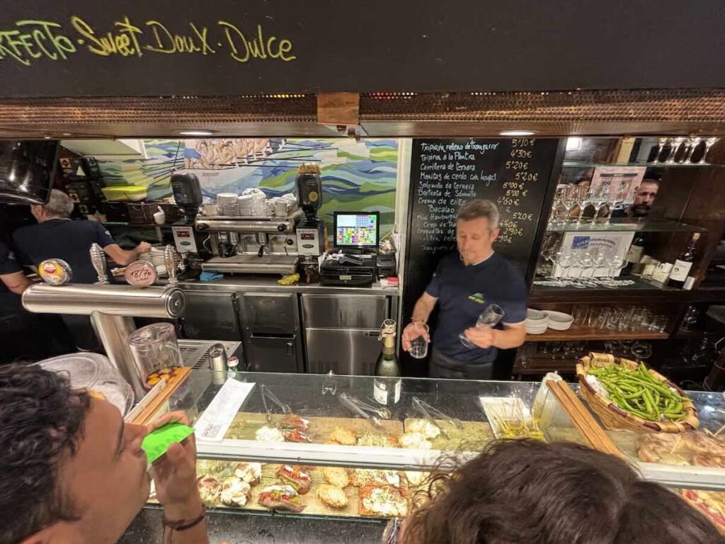Barman serving behind a pintxos counter, with a chalkboard listing hot dishes and prices and a basket of green peppers