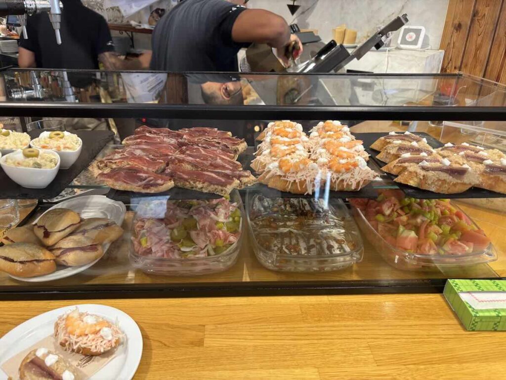 Counter display of pintxos with jamón, prawn, anchovy and tomato toasts on slate trays, and cold dishes in the case below