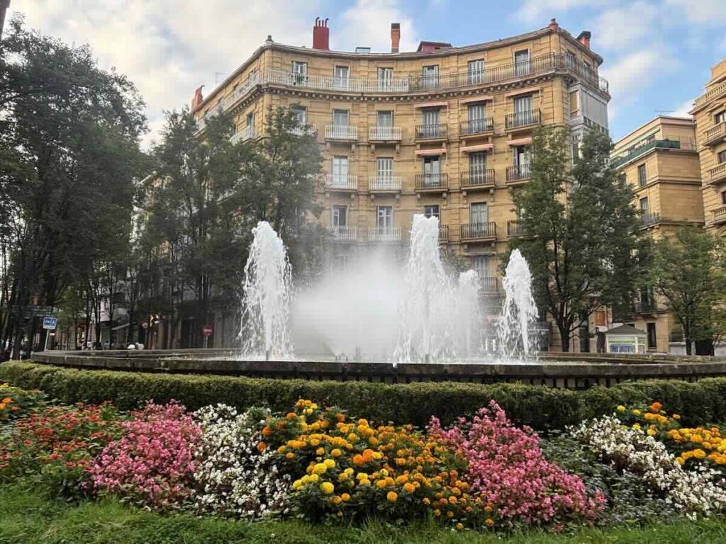 A fountain throwing up three plumes of water in front of a curved sandstone apartment block, with pink, yellow and white flowerbeds in the foreground.