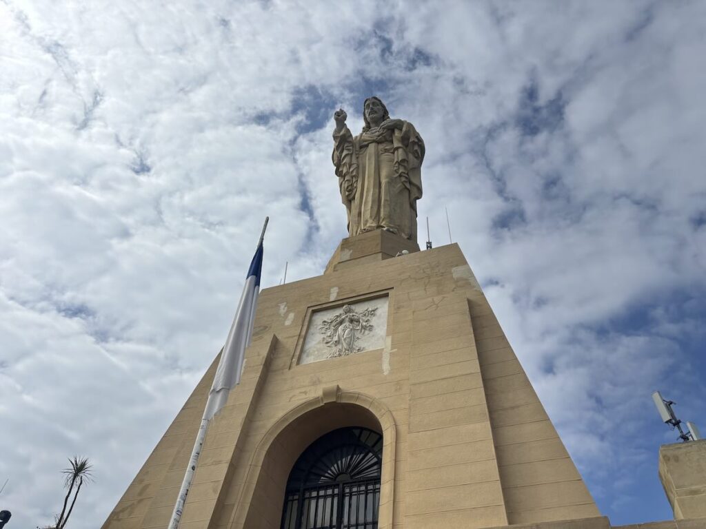 A pale stone statue of Christ with one hand raised, standing on a tall square plinth with an arched gated entrance, photographed from below against broken cloud.
